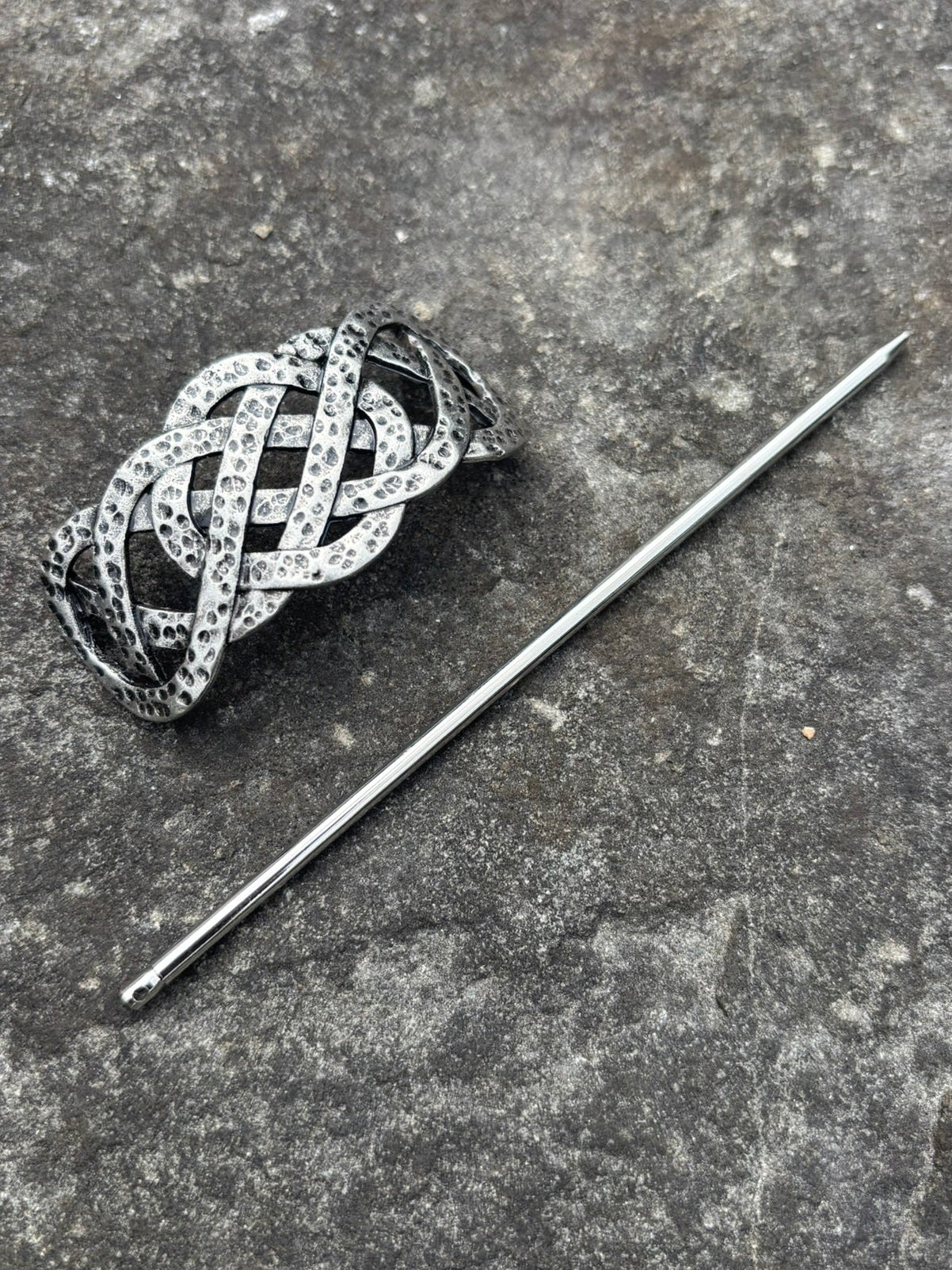 A silver-toned Celtic knot hair slide with small clear stones, displayed next to a silver pin on a textured gray stone surface.