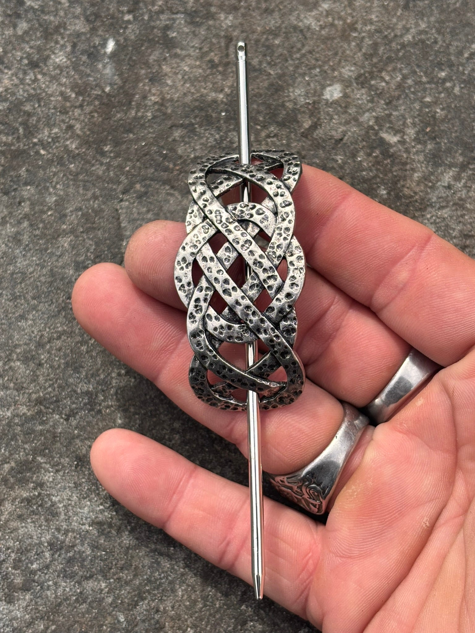A silver-toned Celtic knot brooch held in a hand against a textured gray stone background.