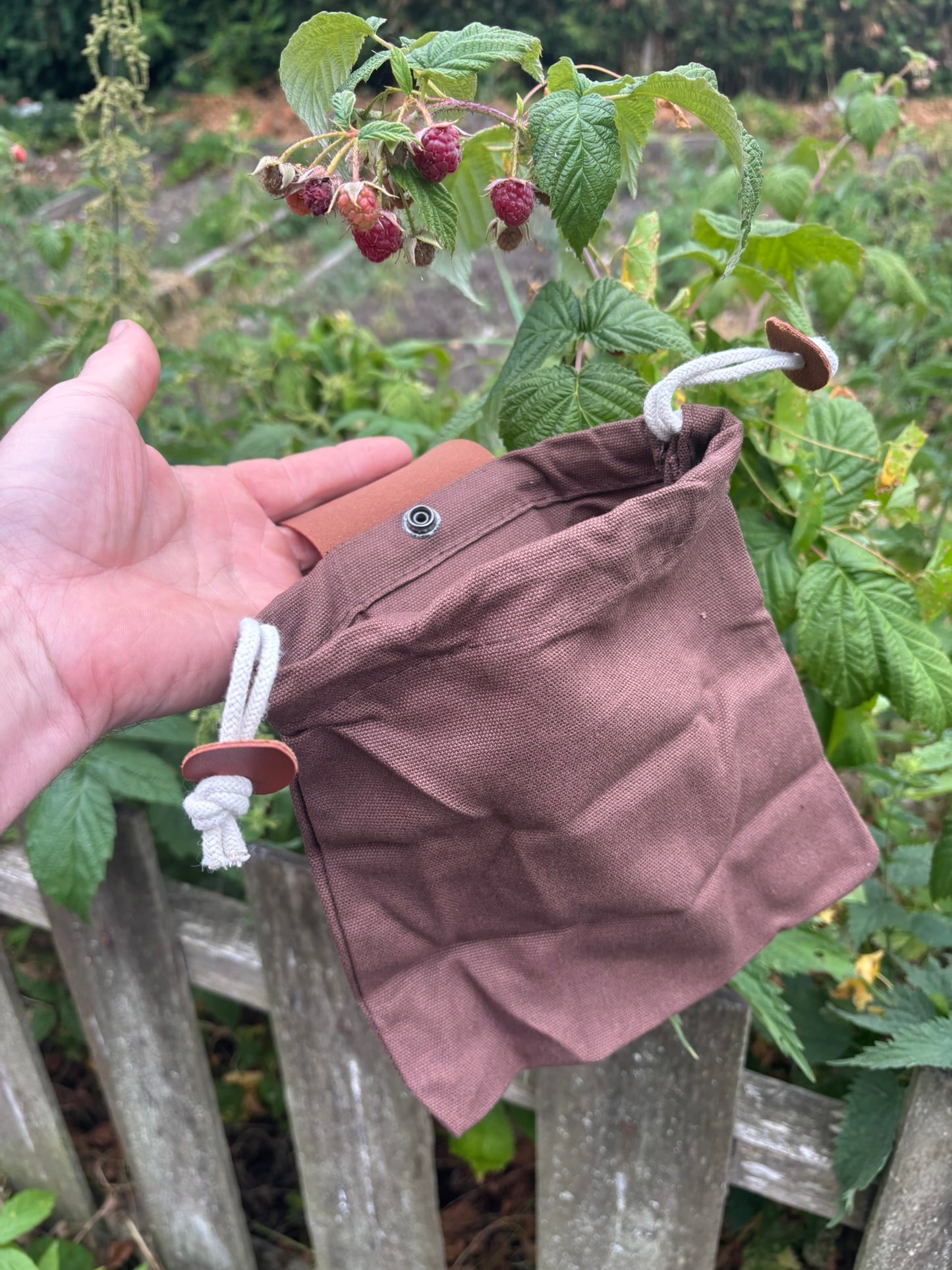 A hand holds a small brown fabric pouch with white drawstrings and a metal snap closure, photographed outdoors in front of green plants and a wooden fence.