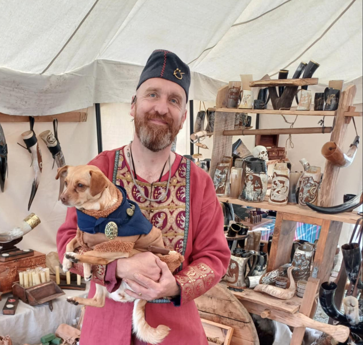 A man wearing a Viking-style costume stands inside a market stall holding a small dog. The costume consists of a dusty rose tunic with ornate gold and green embroidered patterns, a matching dark blue vest, and a black hat.