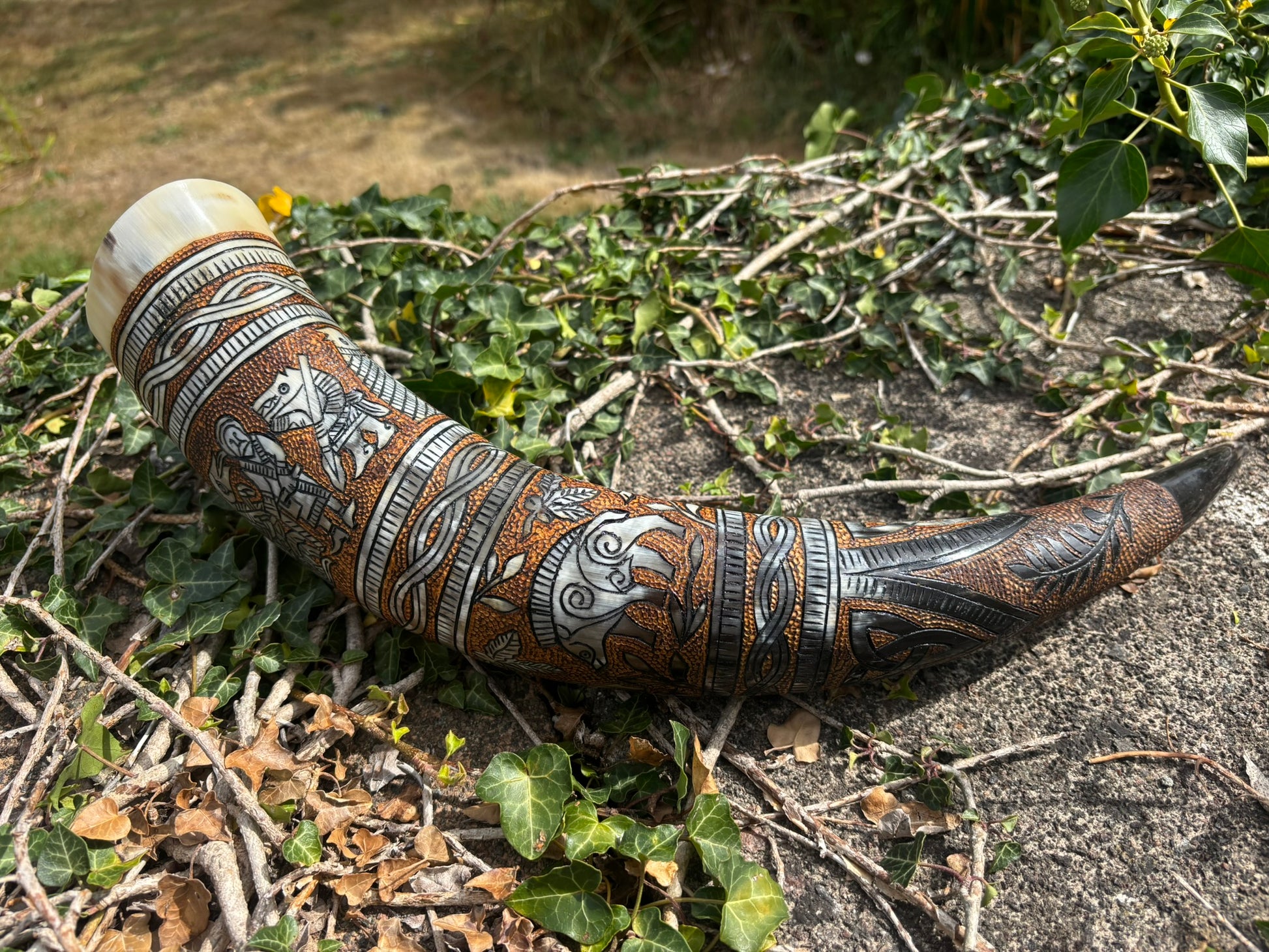 A carved horn or cup with intricate geometric and animal motifs, resting outdoors on a bed of dry leaves, twigs, and green ivy.