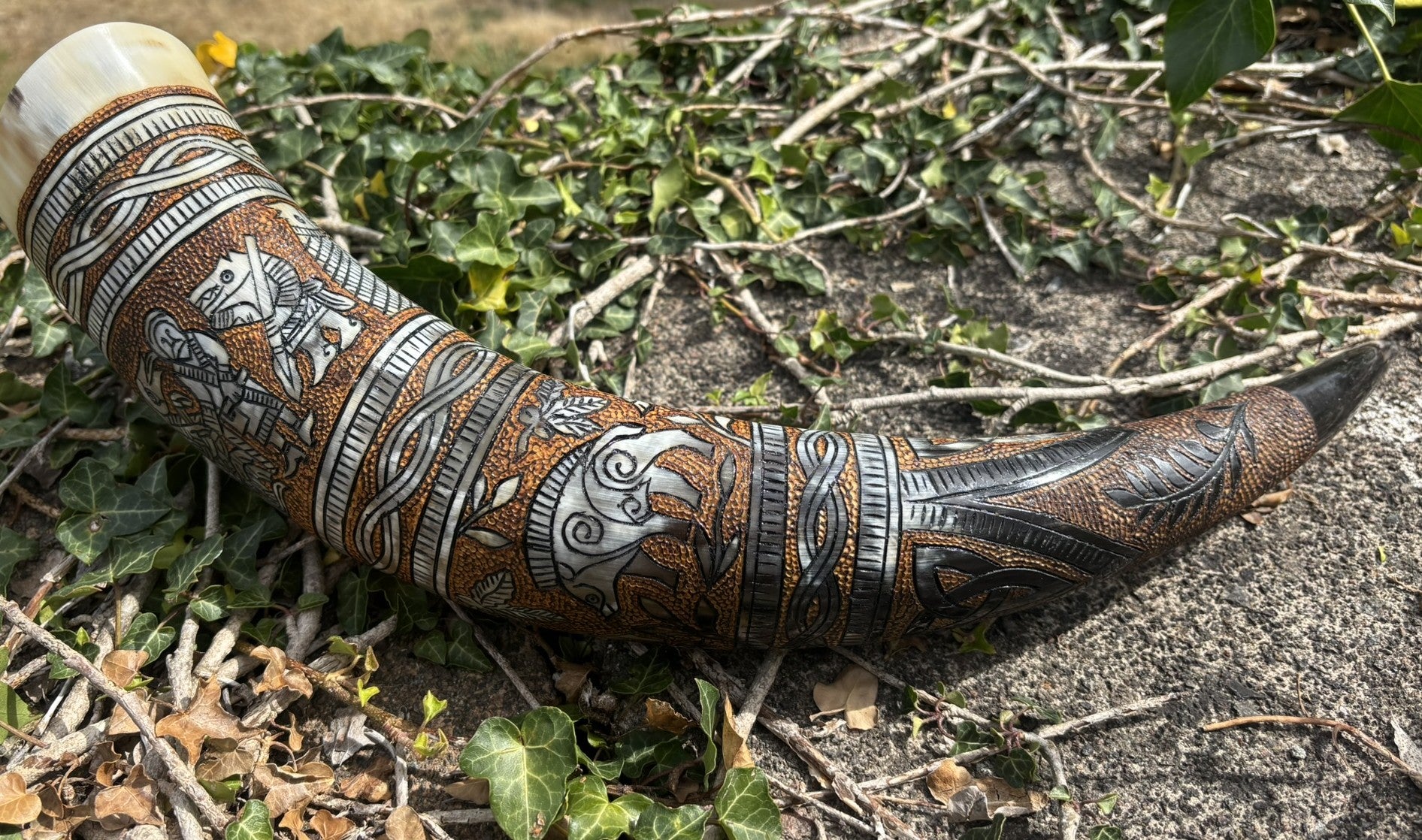 An ornate carved horn or cup with intricate geometric and animal motifs, resting on a natural ground of dirt, twigs, and green ivy.