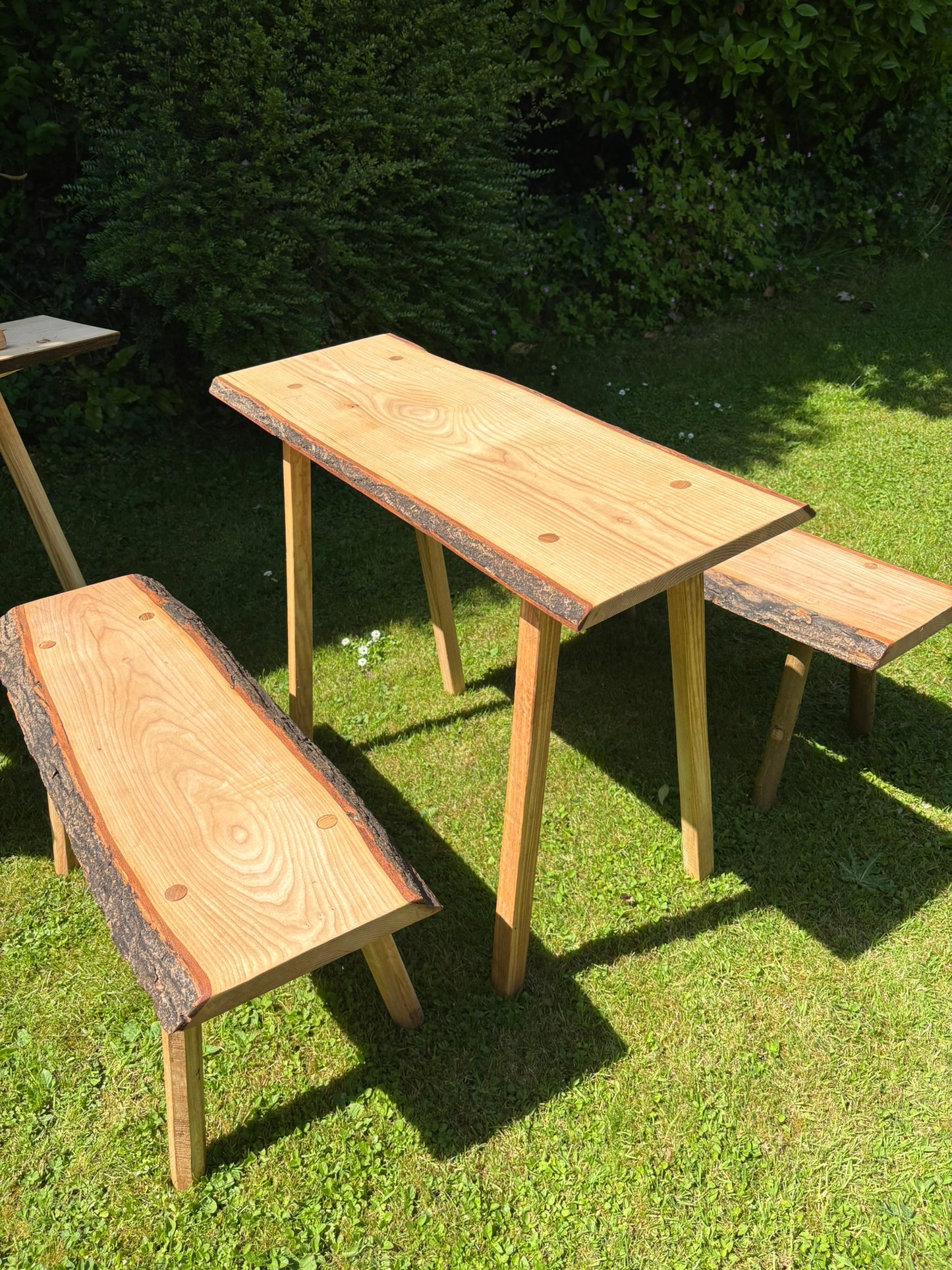 A rustic live-edge wood dining set consisting of a rectangular table and two matching benches, placed on a green grass lawn with a hedge in the background.