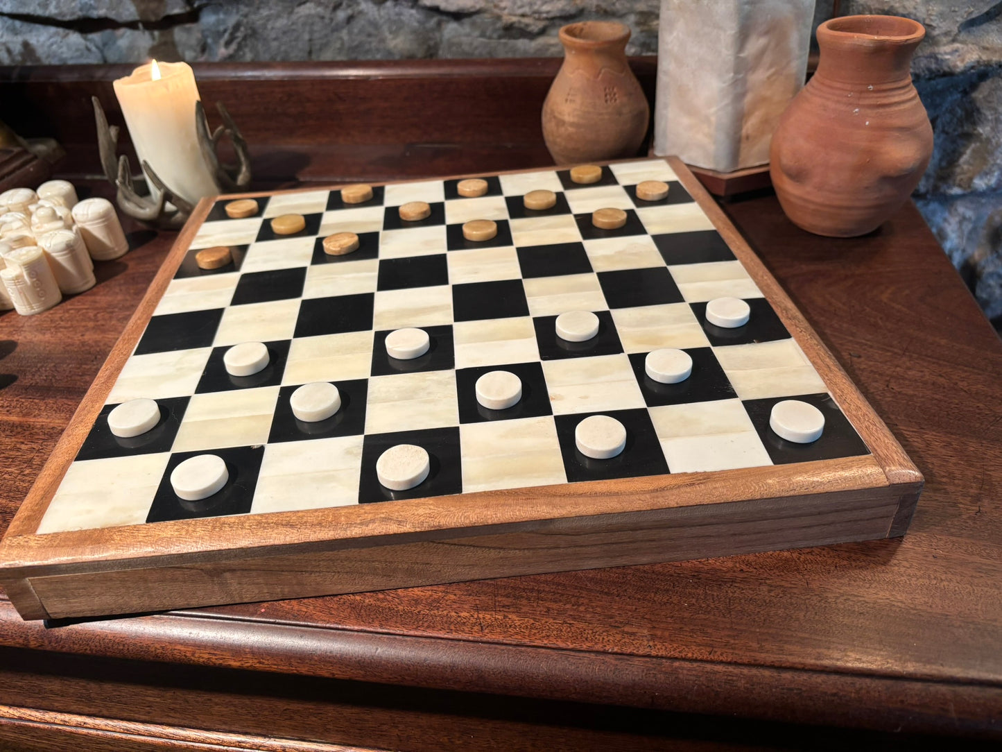 A bone checkers board with black and cream squares, wooden pieces, and a dark wood frame, displayed on a wooden table with decorative vases and a candle in the background.