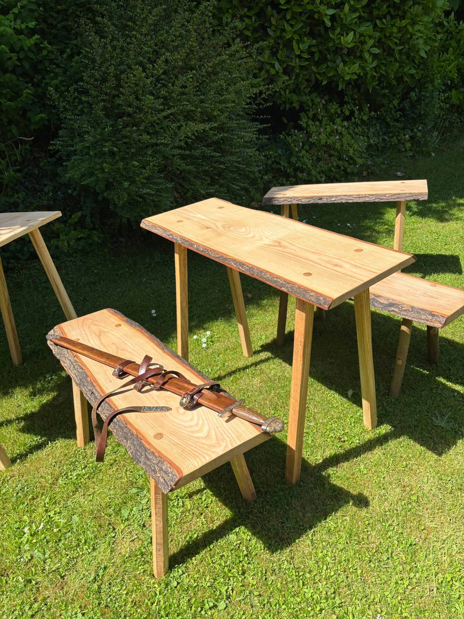 Outdoor wooden dining set with a live-edge table and matching benches on a grassy lawn, with a leather saddle strap draped over one bench.