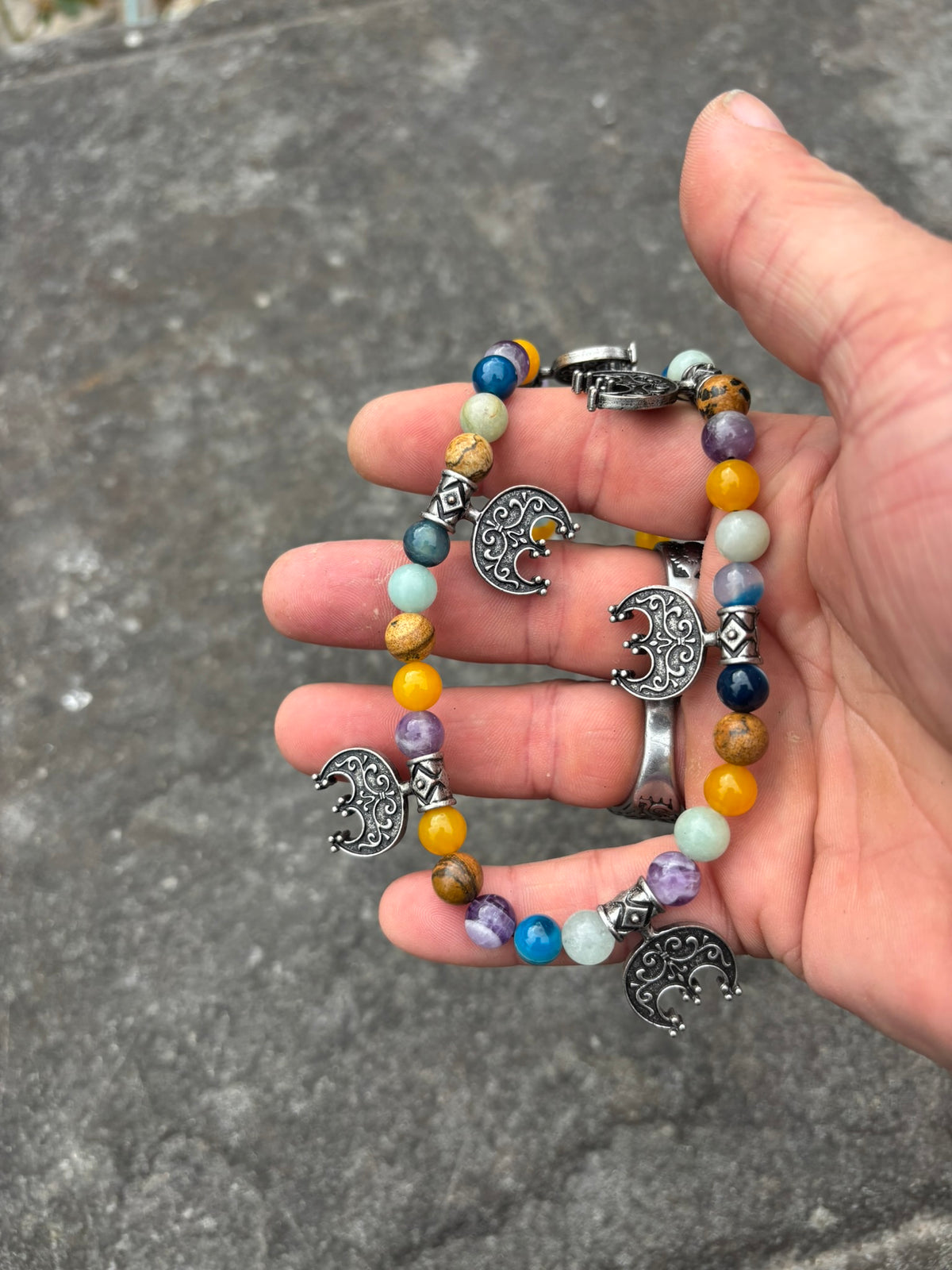 A hand holding a The Moon Maiden's Cord — Viking Lunula & Stone Bead Necklace colorful round beads and silver-toned crescent moon charms, photographed outdoors against a gray stone surface.