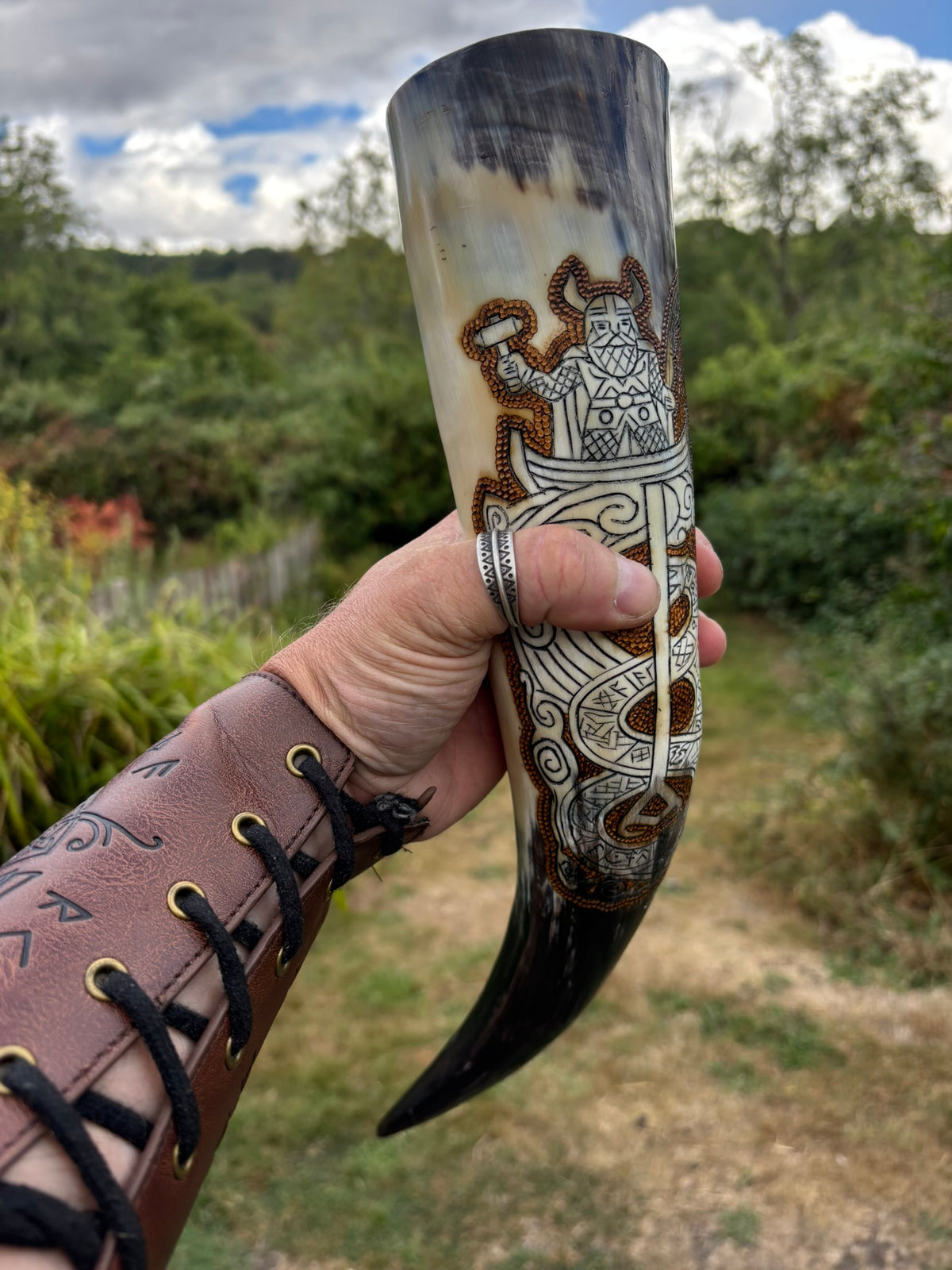A Viking holds a carved horn or shell drinking vessel outdoors, featuring Norse-style runic engravings and a horned figure motif. The vessel has a natural cream-to-dark-brown gradient with blackened edges and detailed white and brown patterns.