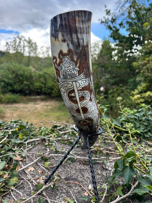 Thor Fishing for Jormungandr Viking Drinking Horn Norse-style runic engravings and a winged motif, displayed on a small tripod stand outdoors among green plants and branches under a cloudy sky.