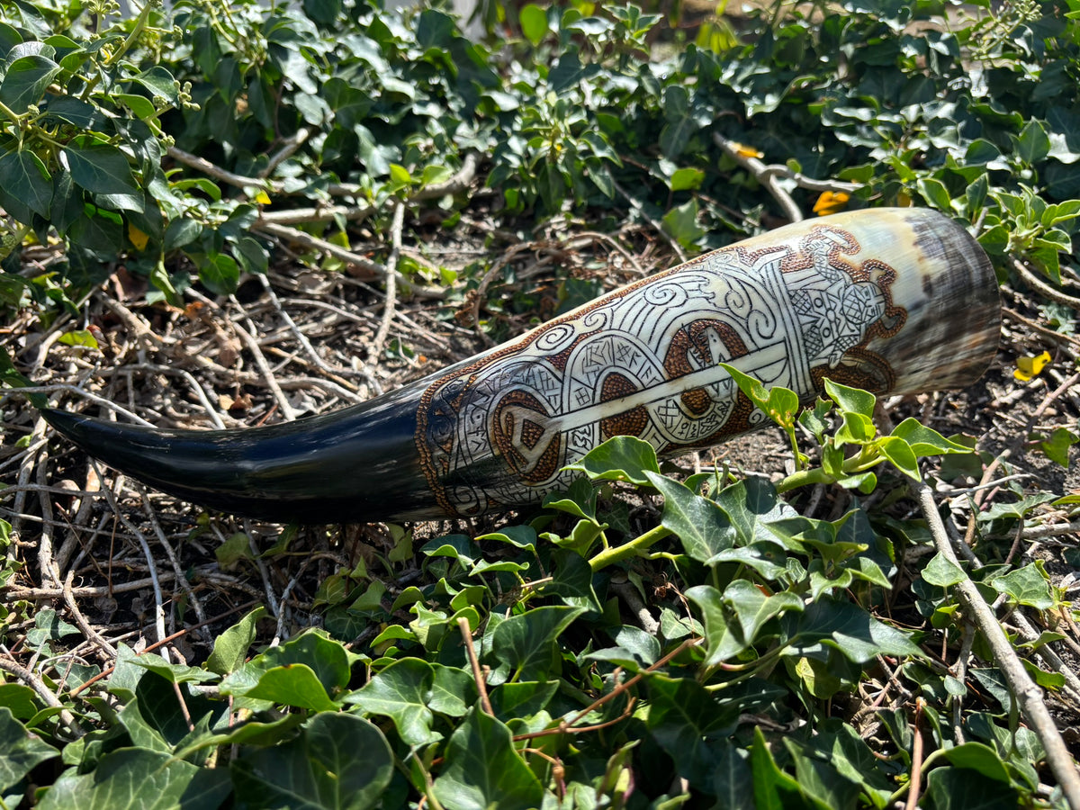 A carved horn or bone decorative object with intricate white and brown geometric patterns, lying on a bed of green ivy and dry twigs outdoors.