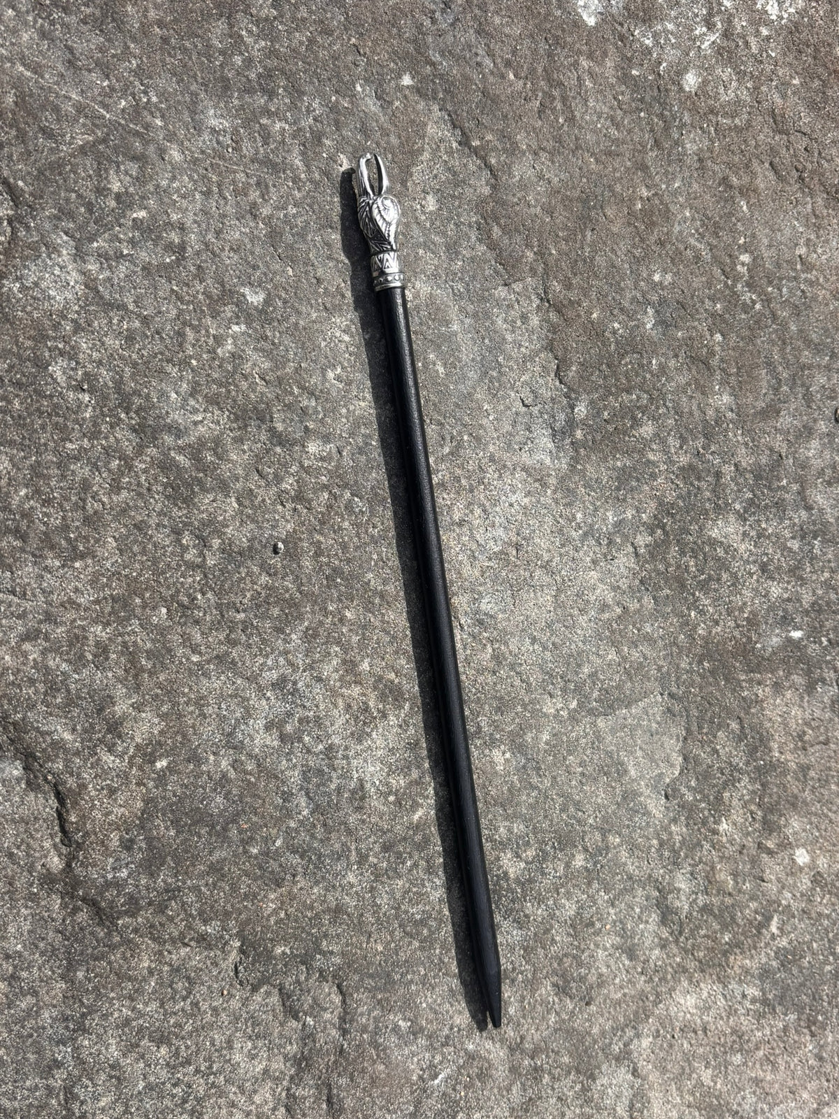 A black hair stick with a silver-toned decorative top, lying diagonally on a textured gray stone surface. Veðrfölnir Eagle Head Hair Pin — Antique Silver
