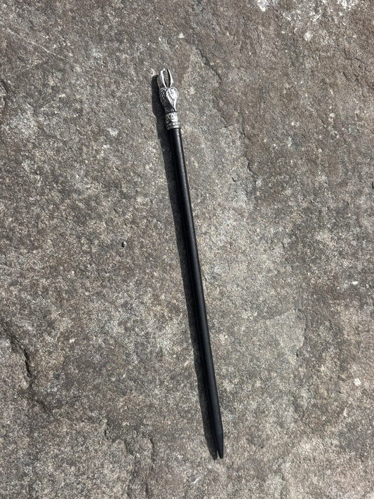 A black hair stick with a silver-toned decorative top, lying diagonally on a textured gray stone surface. Veðrfölnir Eagle Head Hair Pin — Antique Silver