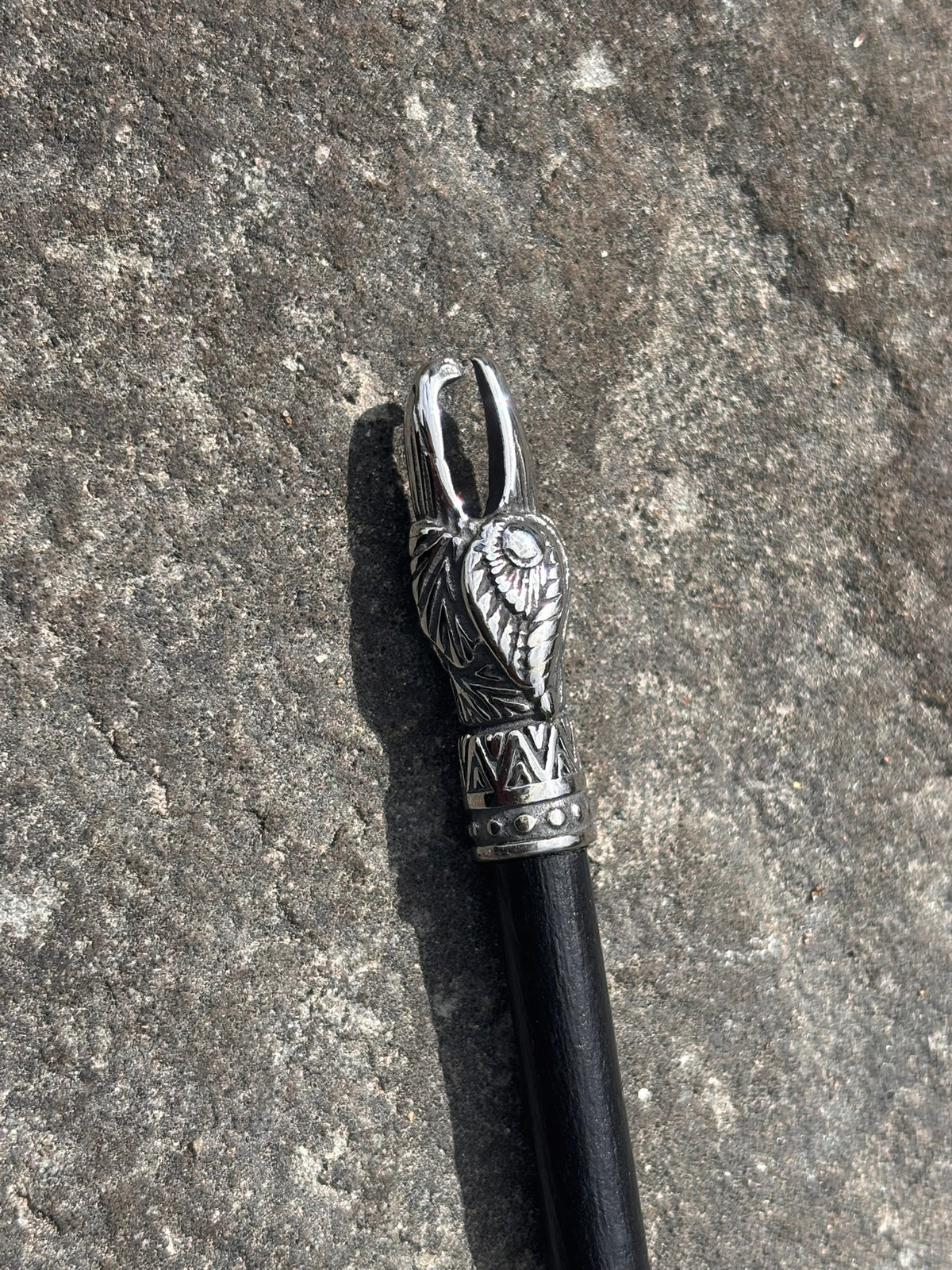 A close-up of a black-handled cane or walking stick with an ornate silver-toned metal top featuring a winged animal head design, placed on a textured gray stone surface.