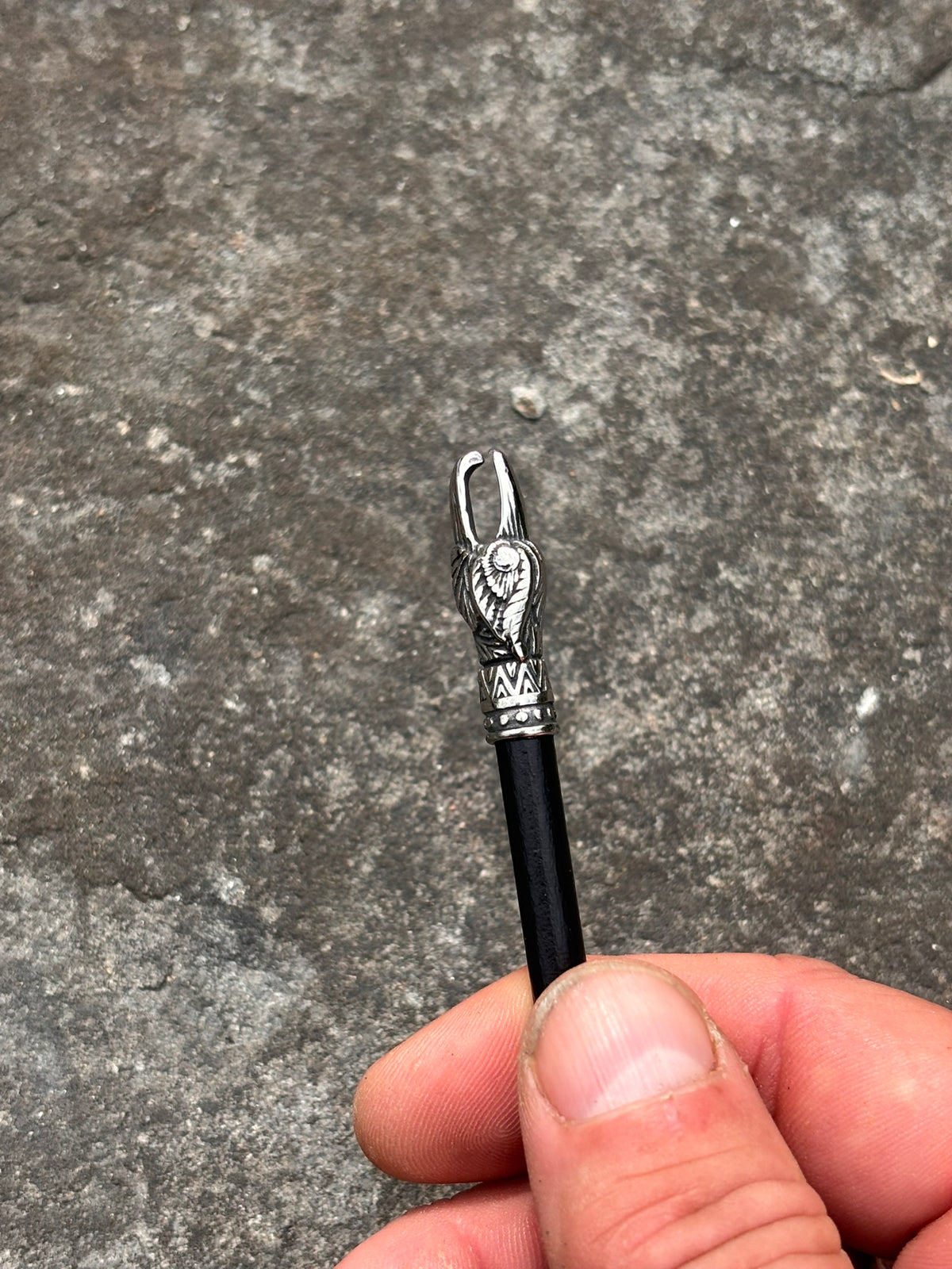 A close-up of a hand holding a black-handled tool with a silver-toned metal looped end featuring an engraved animal head design, set against a textured gray concrete background.