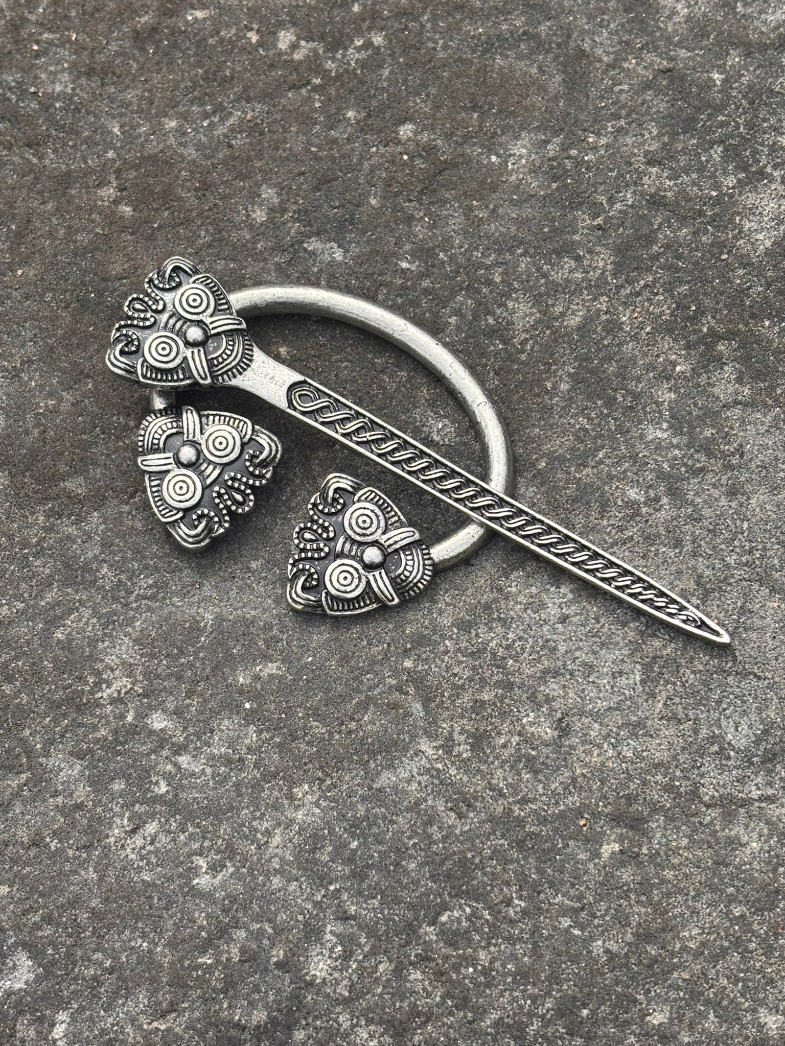 An ornate silver-toned Viking-style brooch with a circular ring, a long engraved blade, and three decorative spiral floral pendants, displayed on a textured gray stone surface.