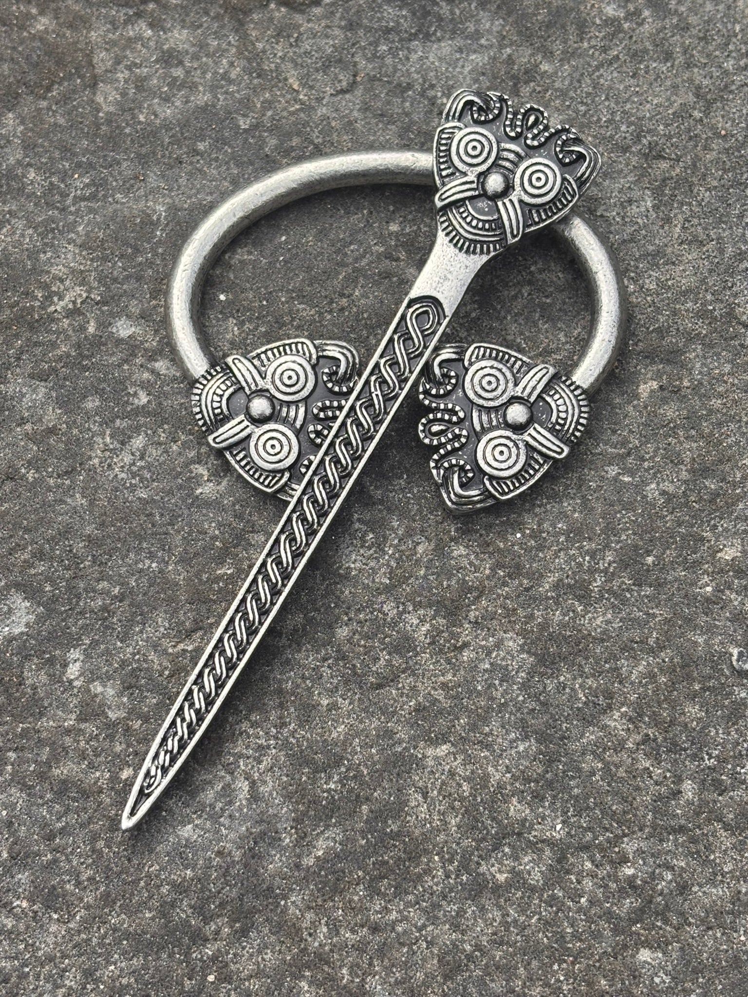 An ornate Borre Cloak Pin, silver-toned metal pin with a circular ring, decorative floral motifs, and a ribbed blade, photographed on a textured gray stone surface.