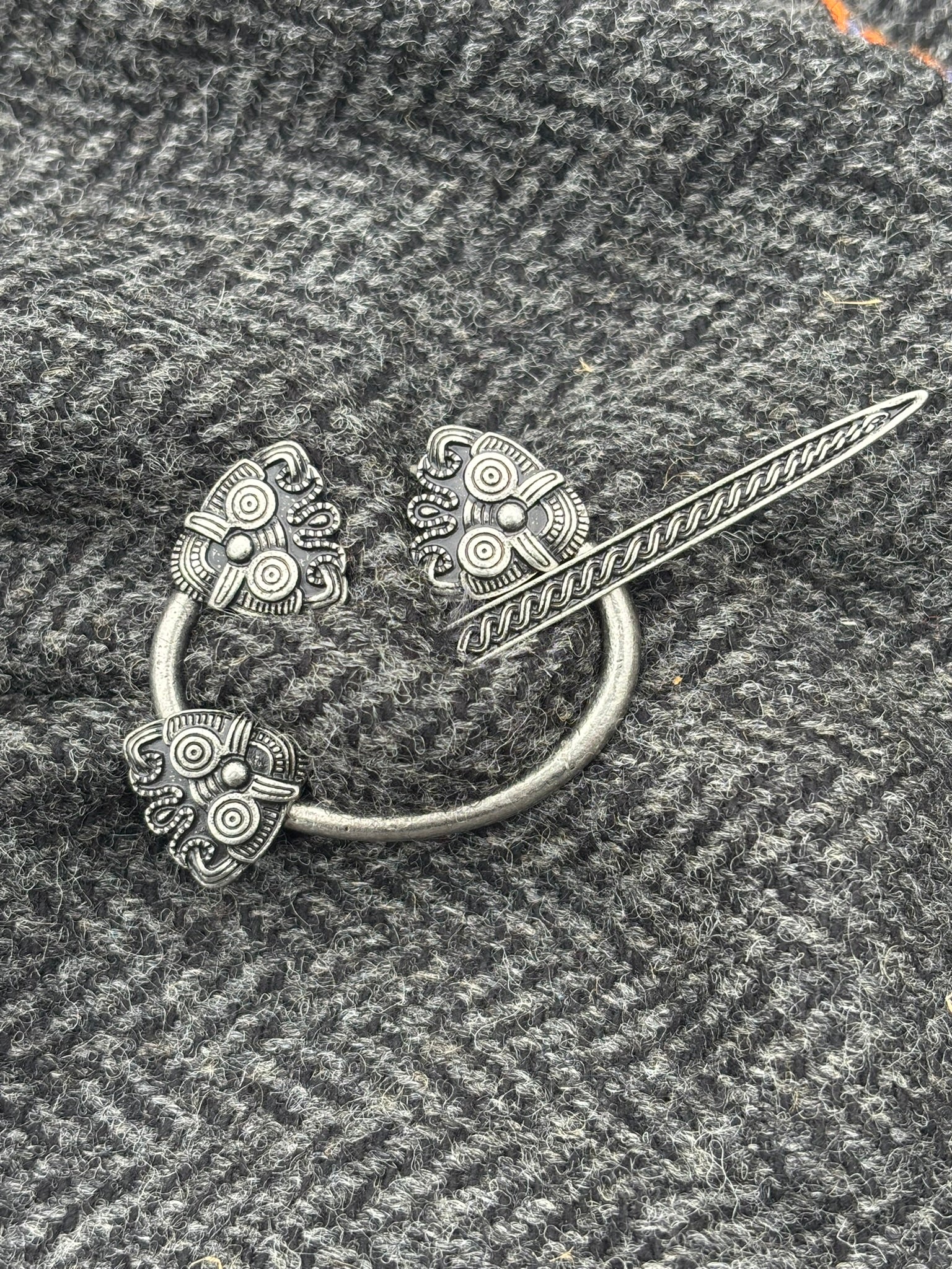 A silver-toned Norse-style ring brooch in Borre style with three ornate, patterned ends, displayed on a textured gray knit fabric.