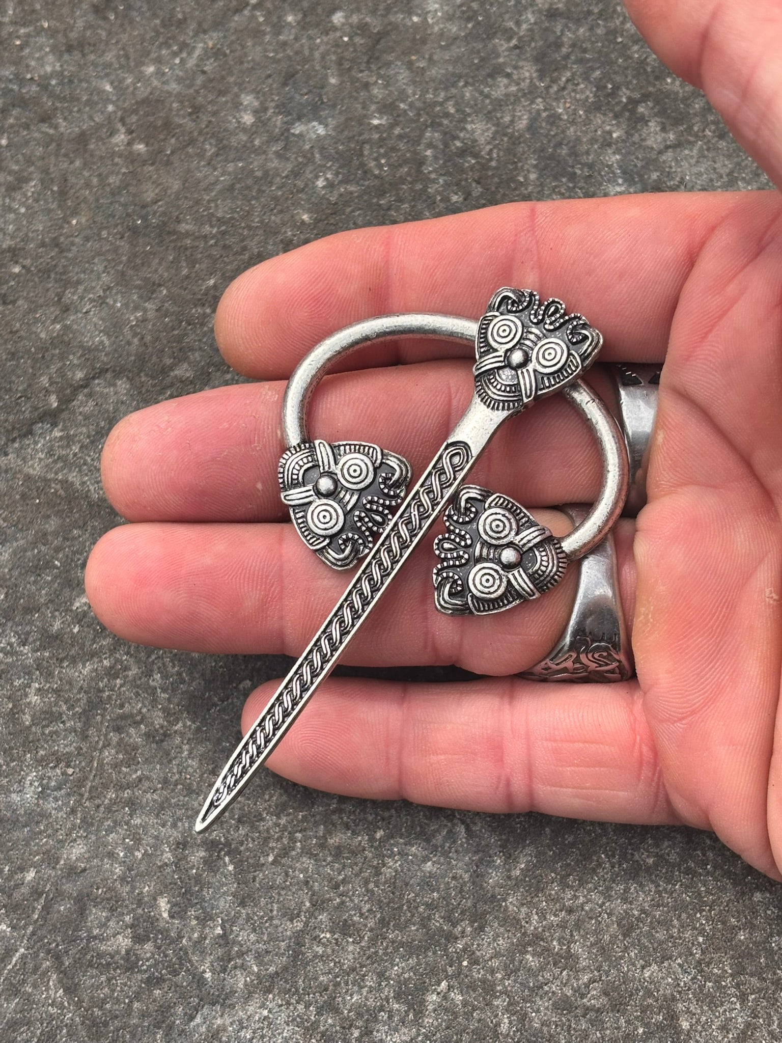 A Viking holding a silver-toned Viking-style ring brooch with a sword motif and ornate knotwork details, set against a textured gray stone background.