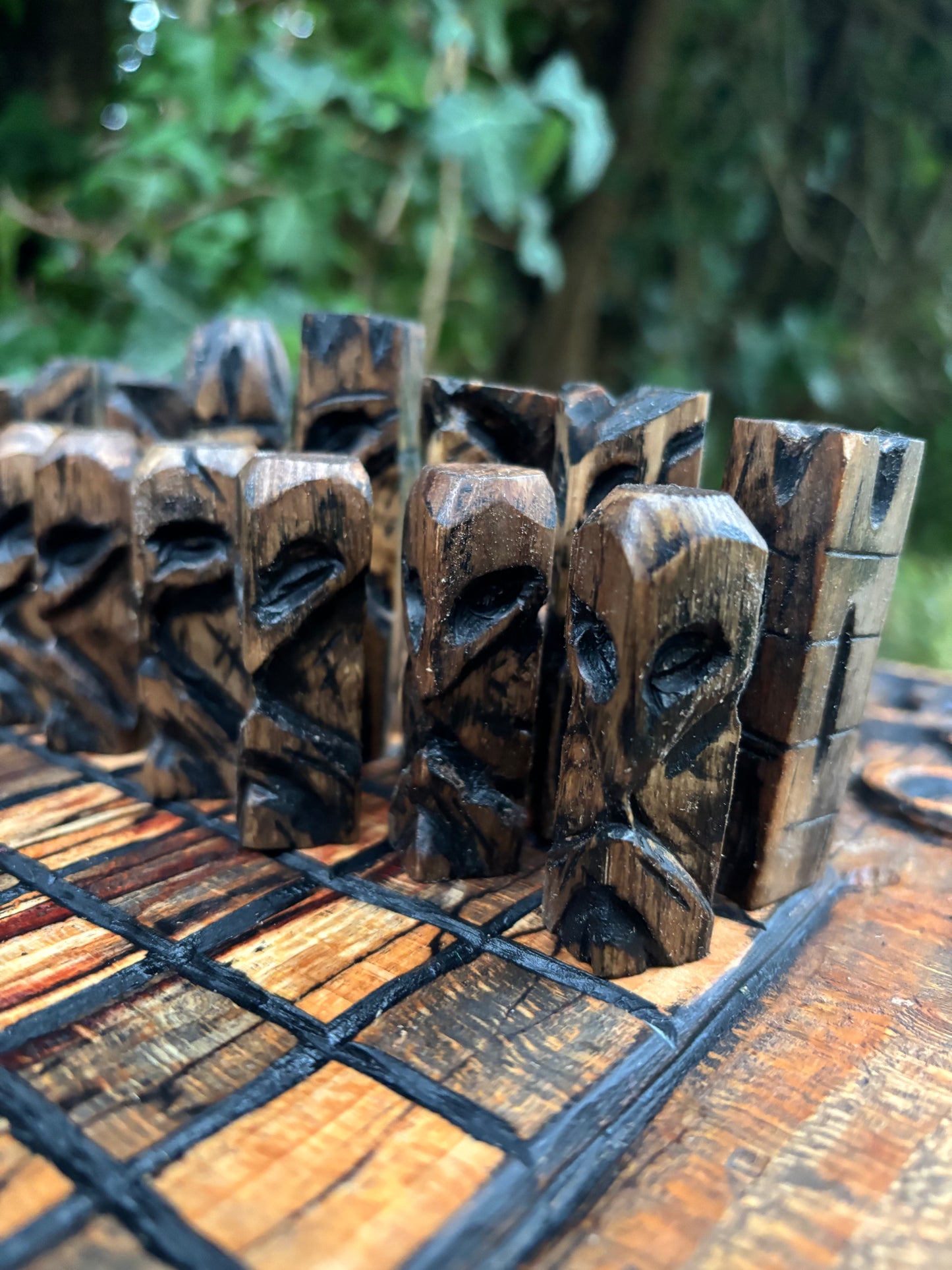 A close-up of carved wooden chess pieces arranged on a wooden board outdoors, with a blurred green forest background.
