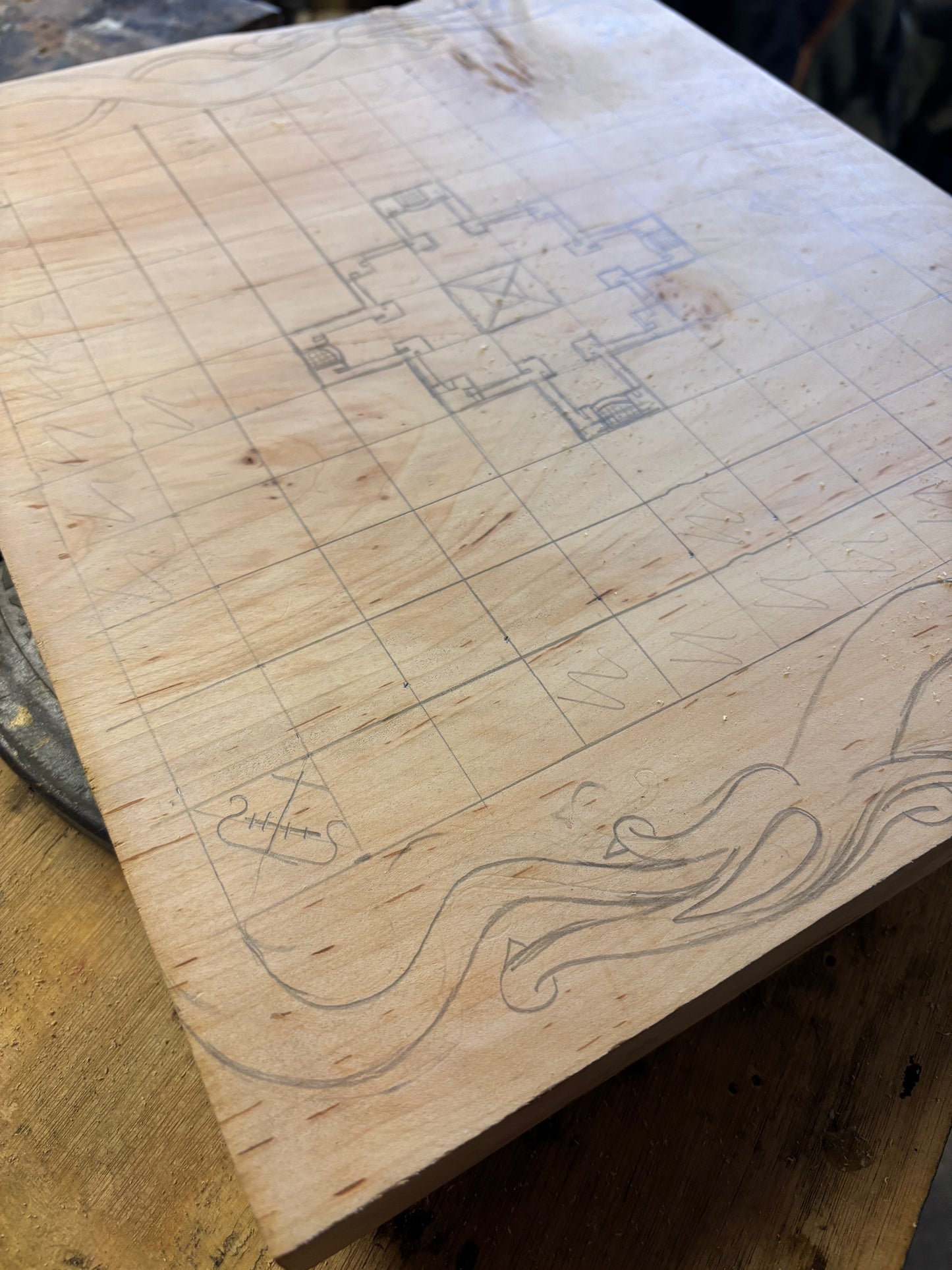 A square unfinished wood board with a hand-drawn grid, a central geometric design, and a wavy line illustration along the bottom edge, photographed on a wooden work surface.