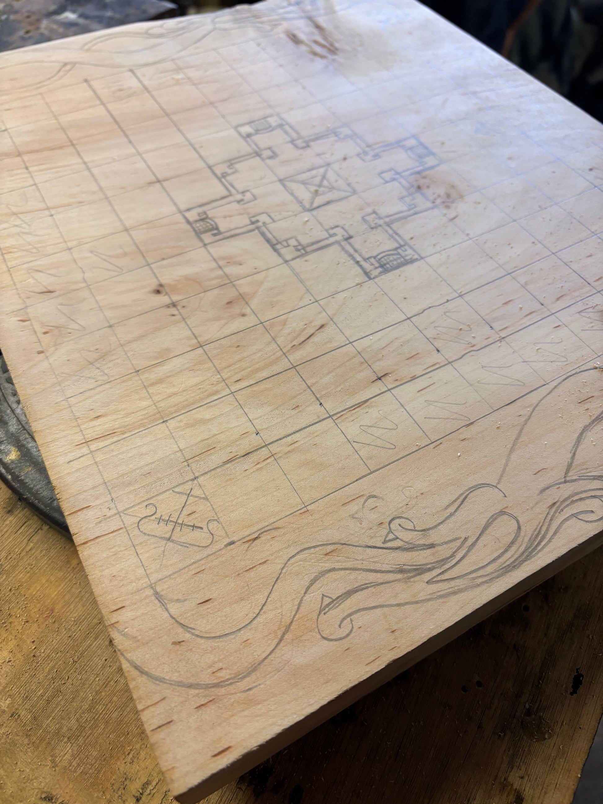 A square unfinished wood board with a hand-drawn grid, a central geometric design, and a wavy line illustration along the bottom edge, photographed on a wooden work surface.