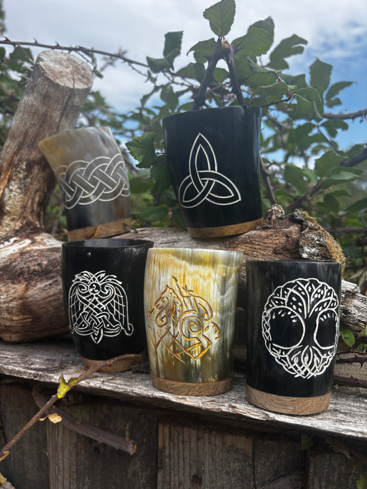 Five handcrafted horn cups with Norse and Celtic designs, displayed outdoors on a rustic wooden surface with logs and green foliage in the background.