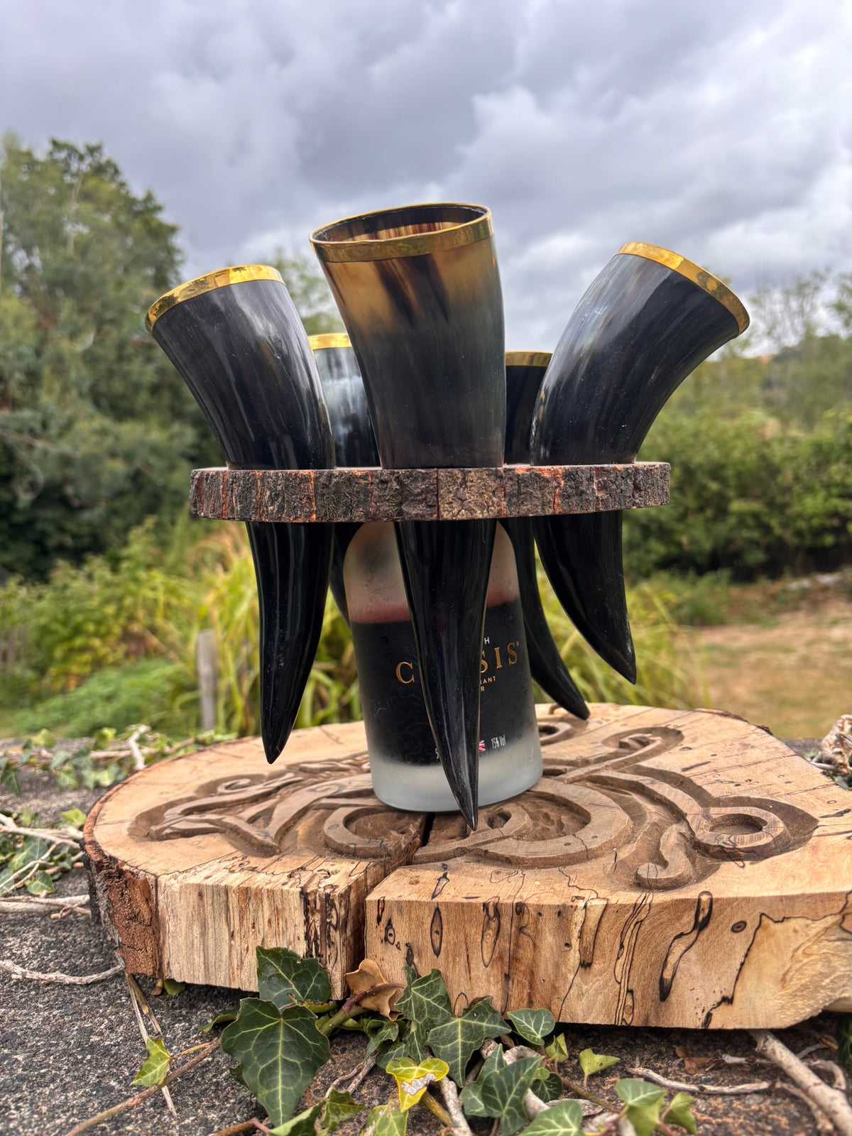 A set of five horn drinking cups with dark brown to black gradient bodies and gold rims, displayed on a carved wooden stand outdoors.