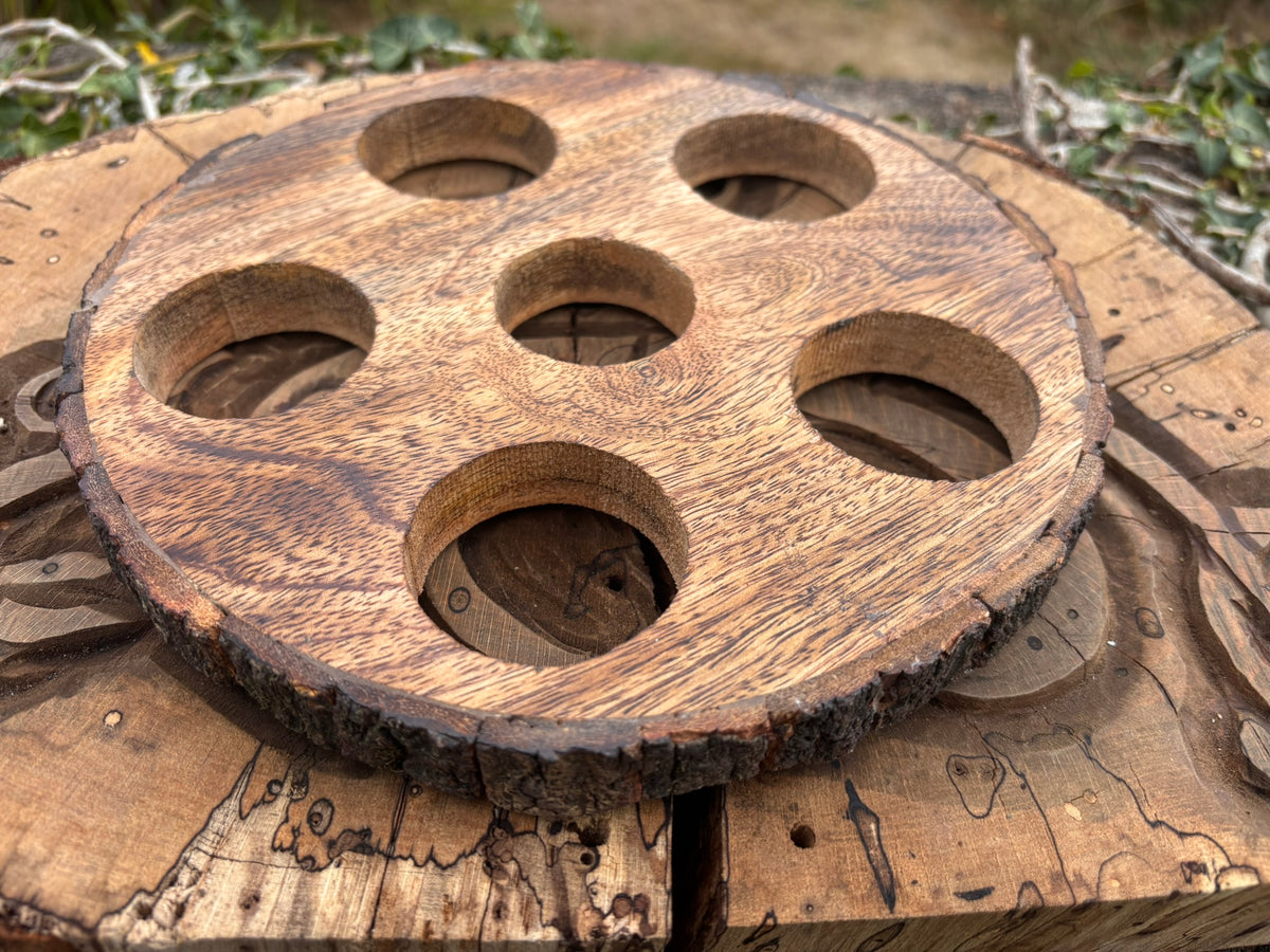 A round wooden wine glass holder or coaster with six circular cutouts, made from a natural wood slice with bark edges, displayed on a rustic wooden surface.