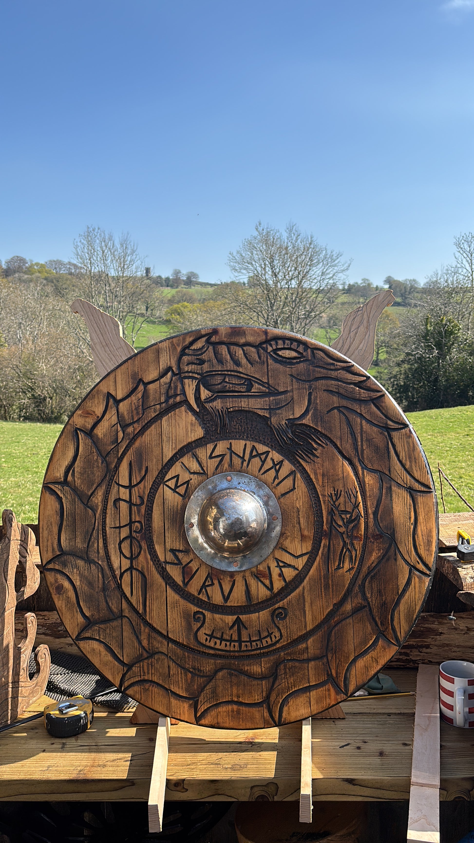 A large hand-carved wooden Viking-style shield with Norse runes and a polished steel bell, displayed outdoors on a wooden stand with a grassy field and trees in the background.