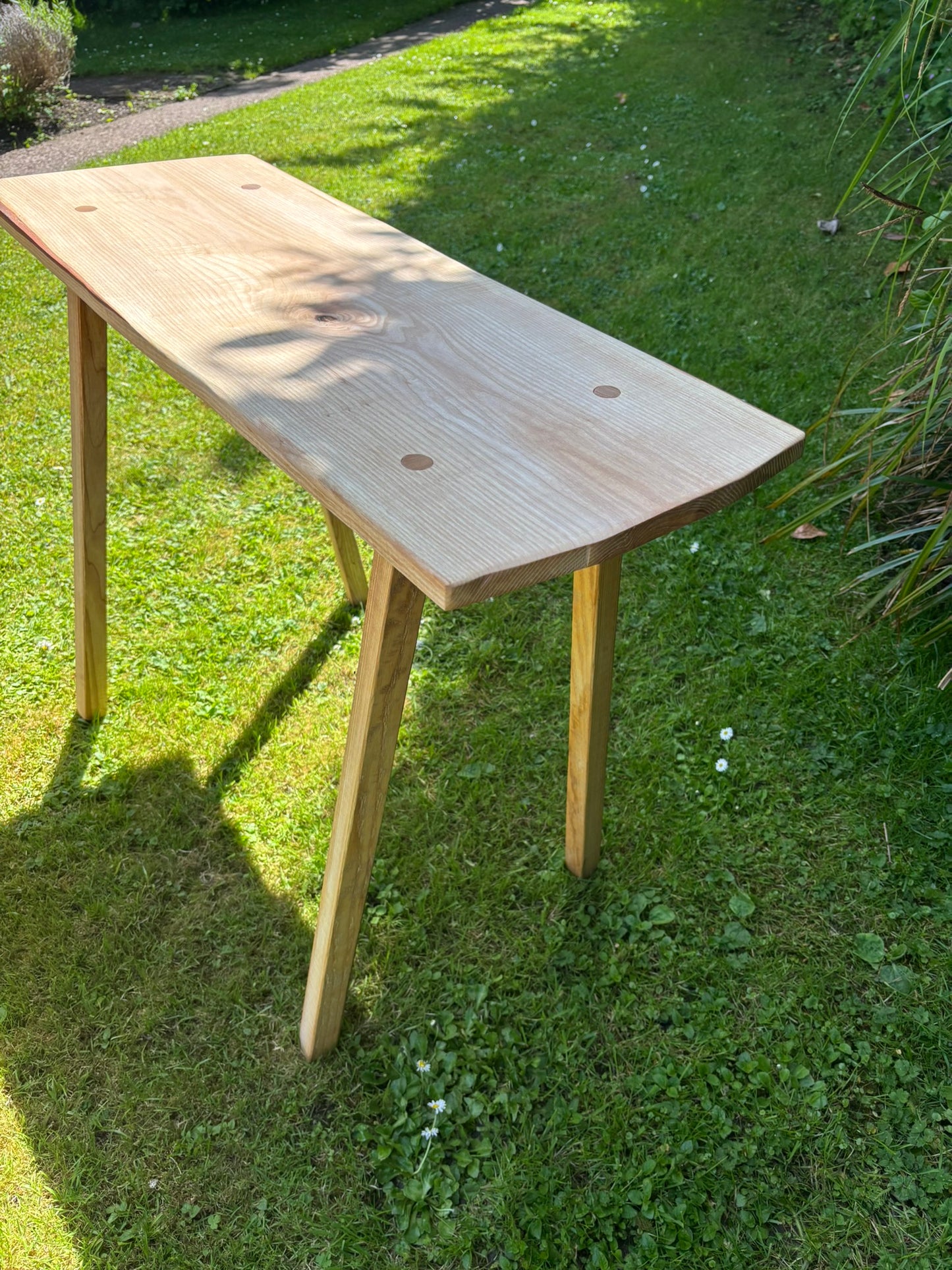 A light natural wood console table with a live edge and three legs, photographed outdoors on a green grass lawn in bright daylight.