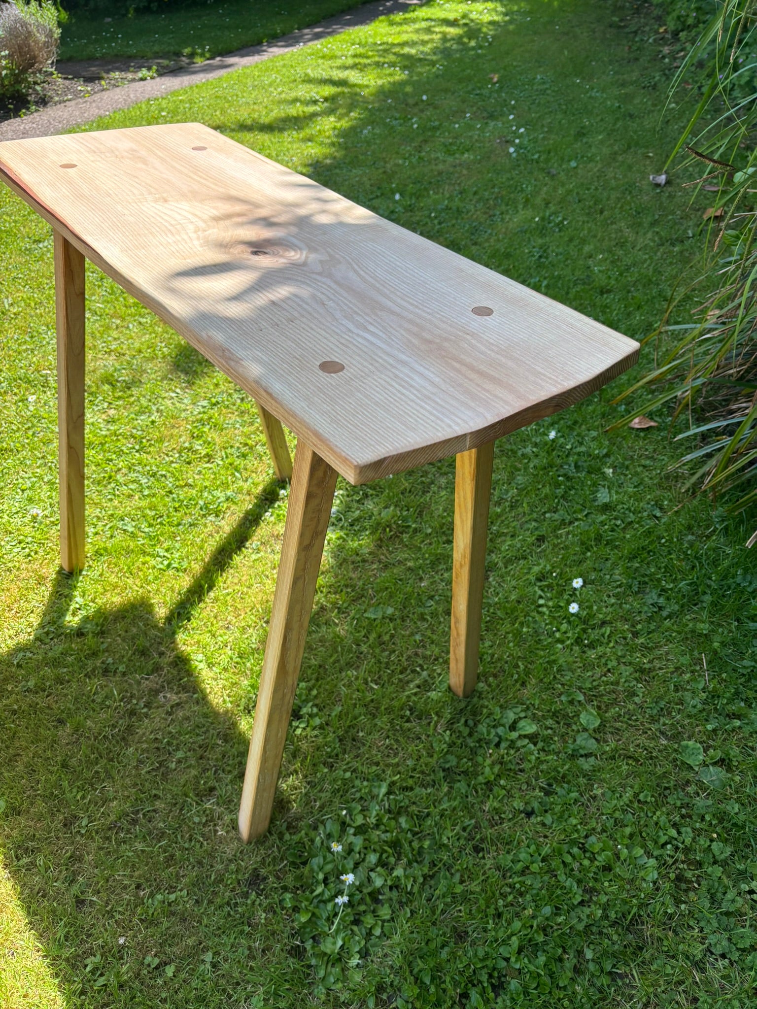 A light natural wood console table with a live edge and three legs, photographed outdoors on a green grass lawn in bright daylight.