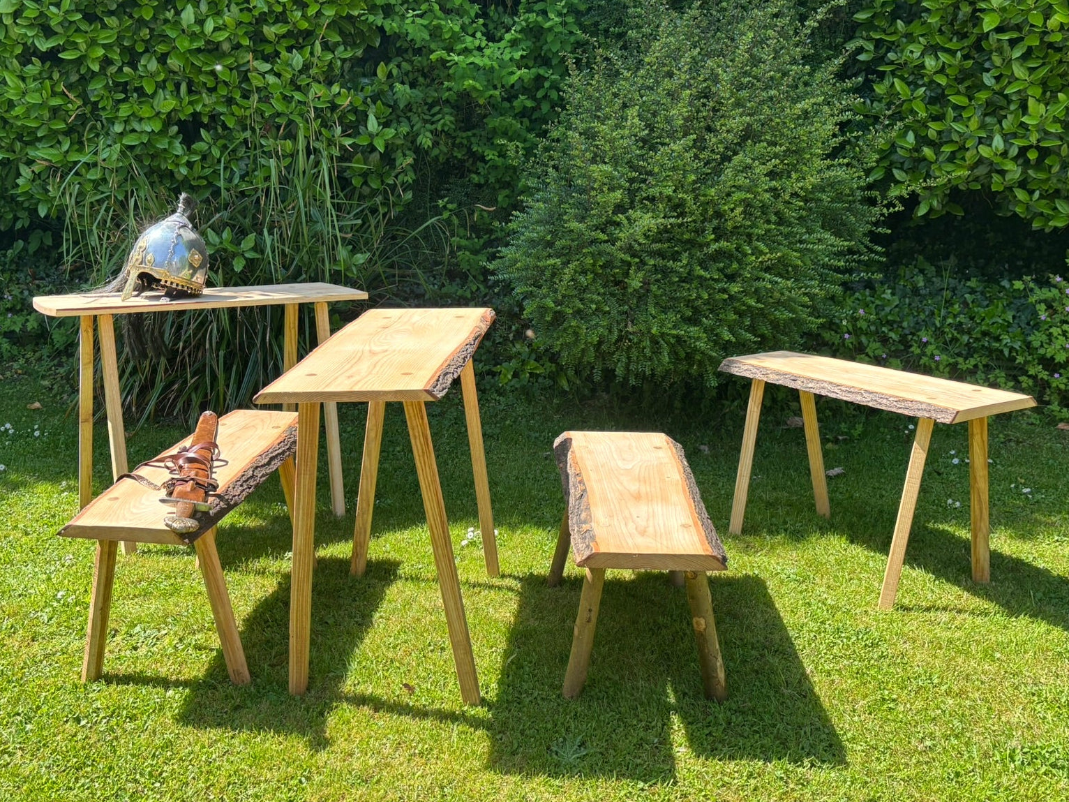A set of five rustic wooden outdoor furniture pieces, including three tables and two benches, arranged on a green lawn with a dense green hedge in the background.