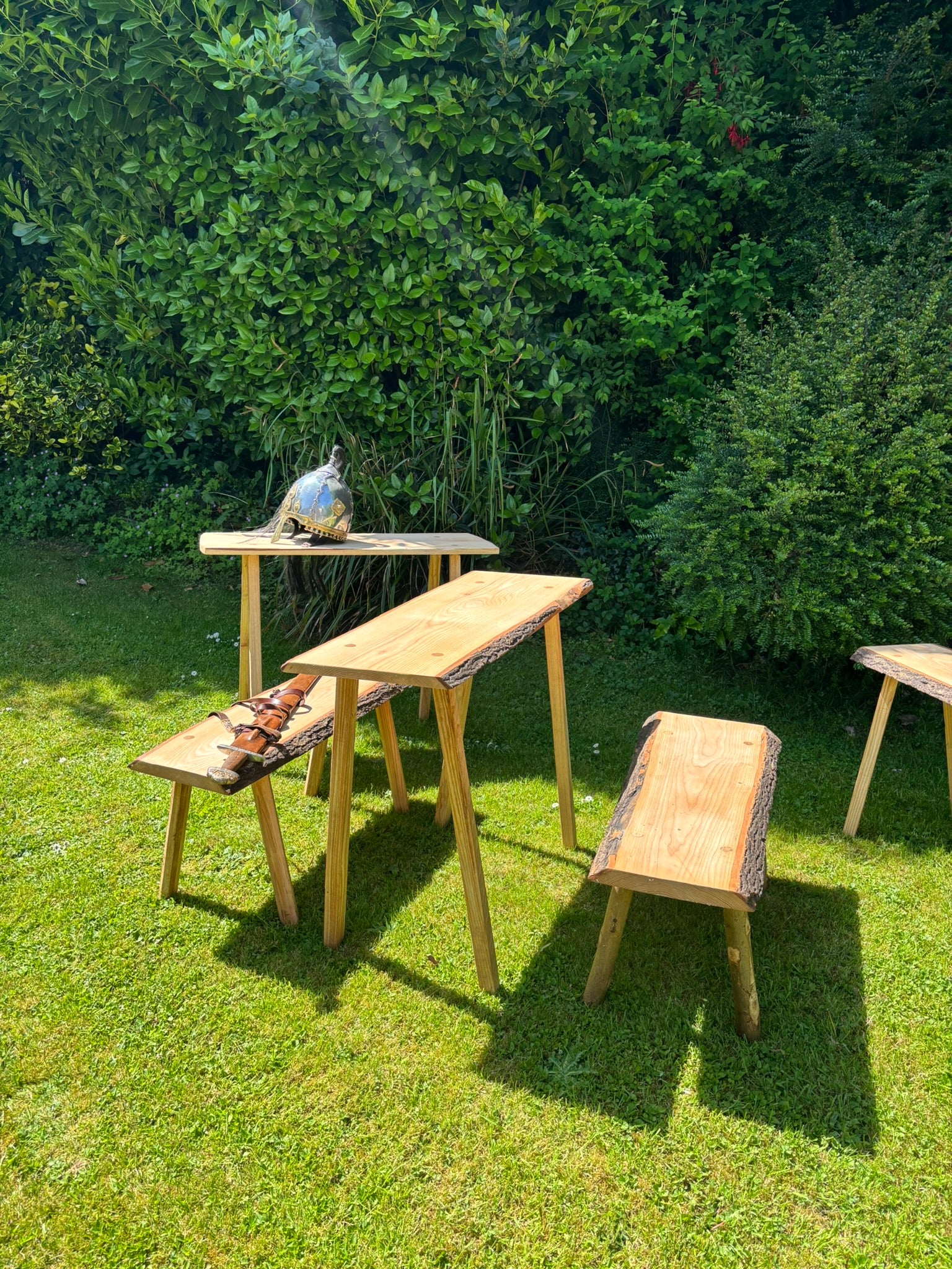 Outdoor wooden Viking dining set with a table and benches on a grassy lawn in front of a dense green hedge. The set includes a rectangular table with a live-edge wood top and matching benches, with a small decorative bird figurine on one of the tabletops.