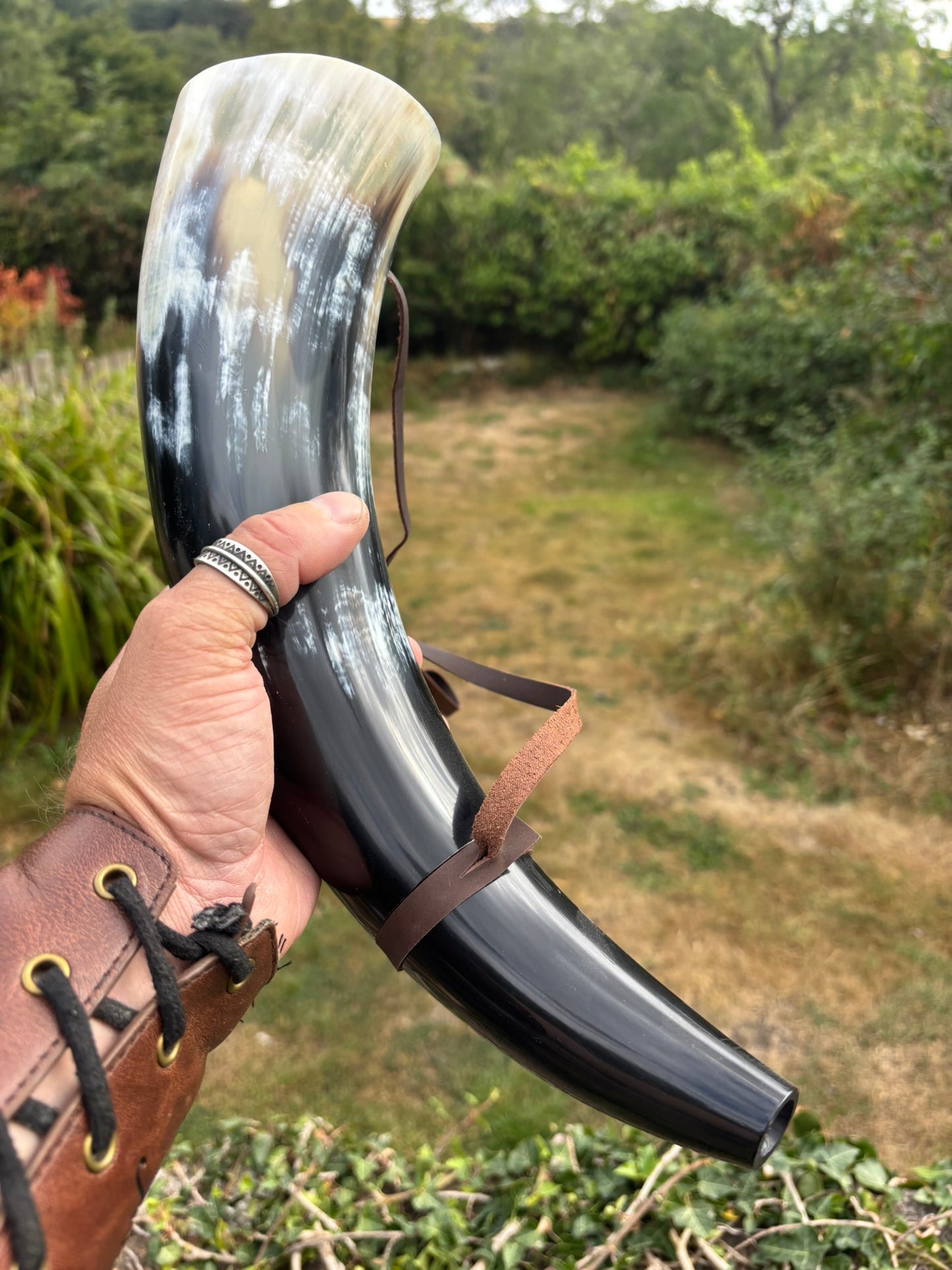 A Viking holding a war horn outdoors, with a brown leather strap around the base. The background shows a blurred garden or wooded outdoor setting.