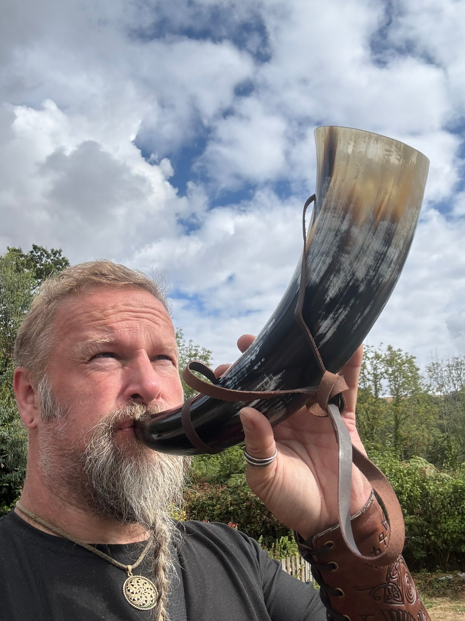 A bearded man outdoors drinking from a large horn-shaped vessel with a brown leather strap, set against a cloudy blue sky and green trees.