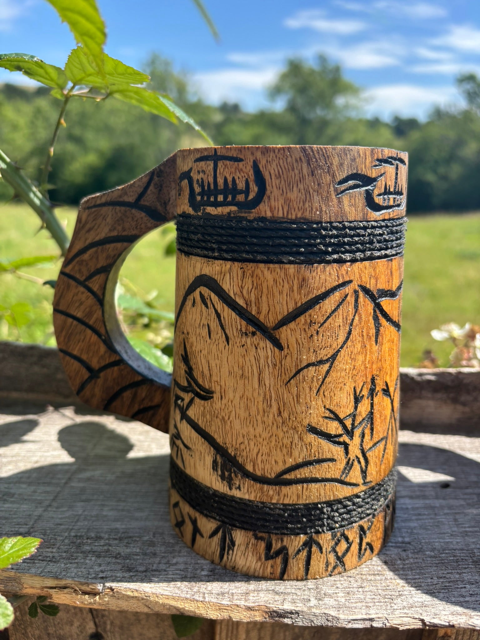 A carved wooden mug with a handle, featuring Norse-style runic symbols and mountain illustrations, set outdoors on a rustic wooden surface with a blurred green field and blue sky in the background.