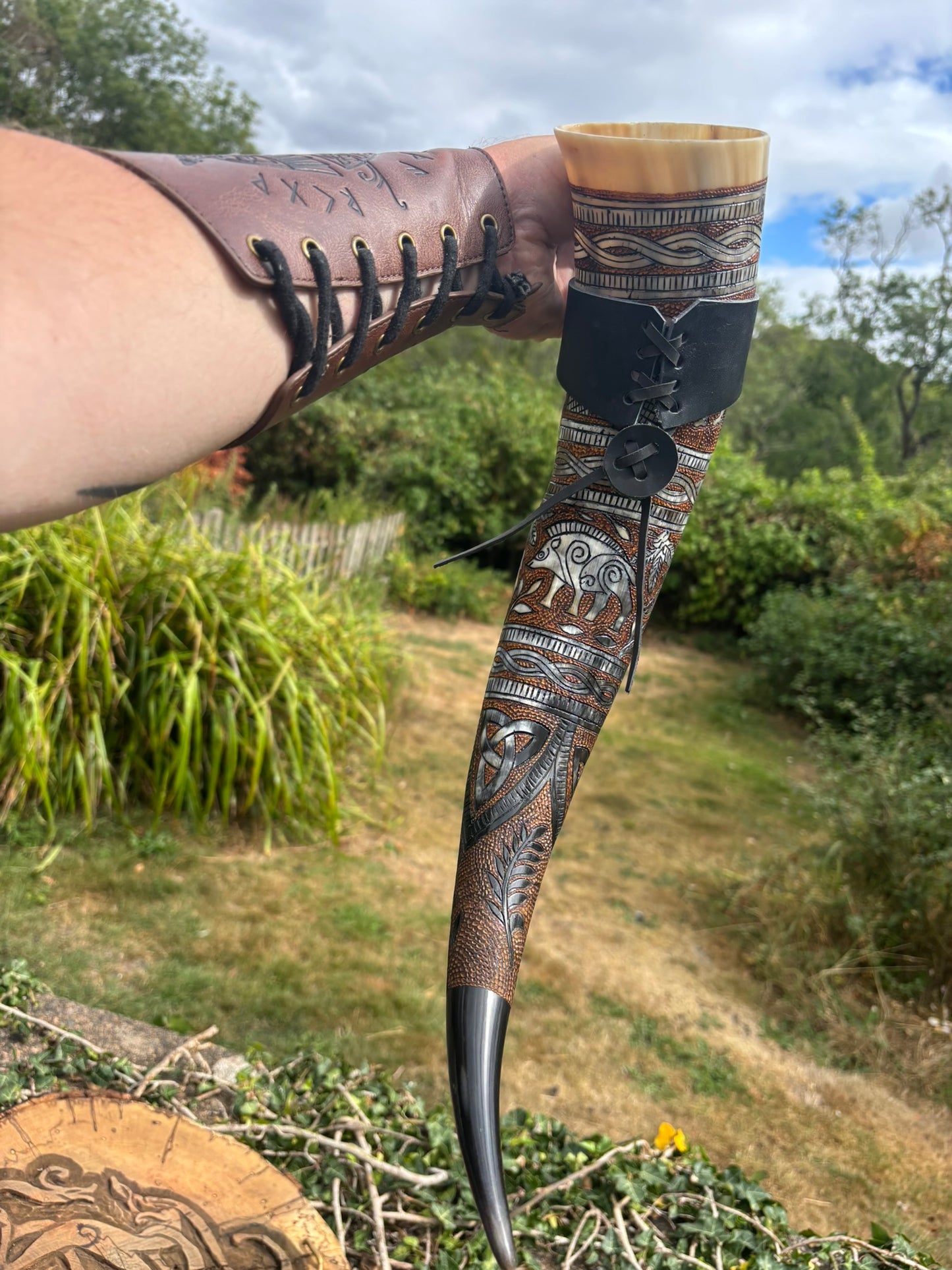 A hand holds a carved horn or bone pipe against an outdoor garden background. The pipe has intricate geometric and animal-style engravings in dark brown, black, and tan, with a dark brown leather wrap and laces on the upper portion and a black metal tip.