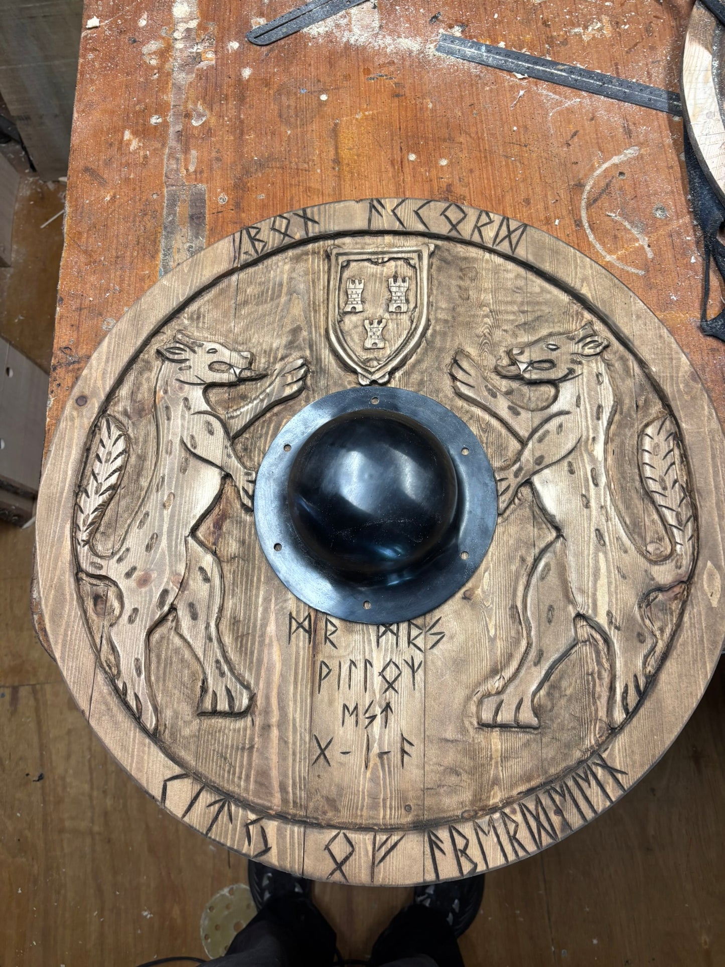 A round wooden medieval-style shield with carved animal figures, runic inscriptions, and a central black metal boss, photographed on a wooden workbench.